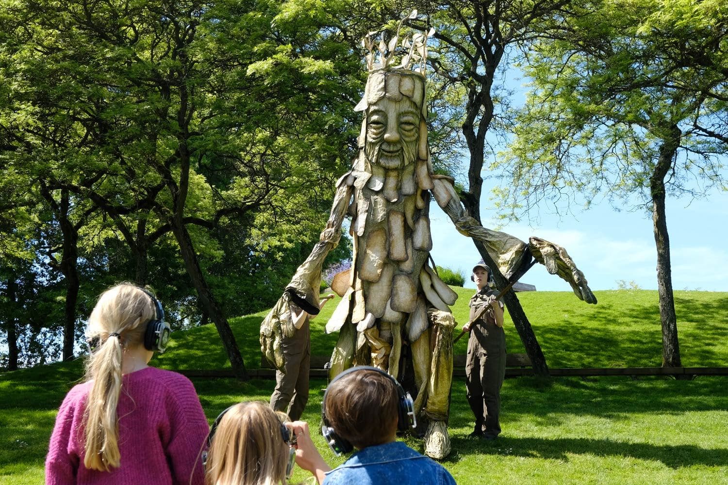 A 5-metre tall tree puppet with a smiling face being performed by five puppeteers on a grassy hill lined with trees. 3 kids wearing headphones watch the performance.