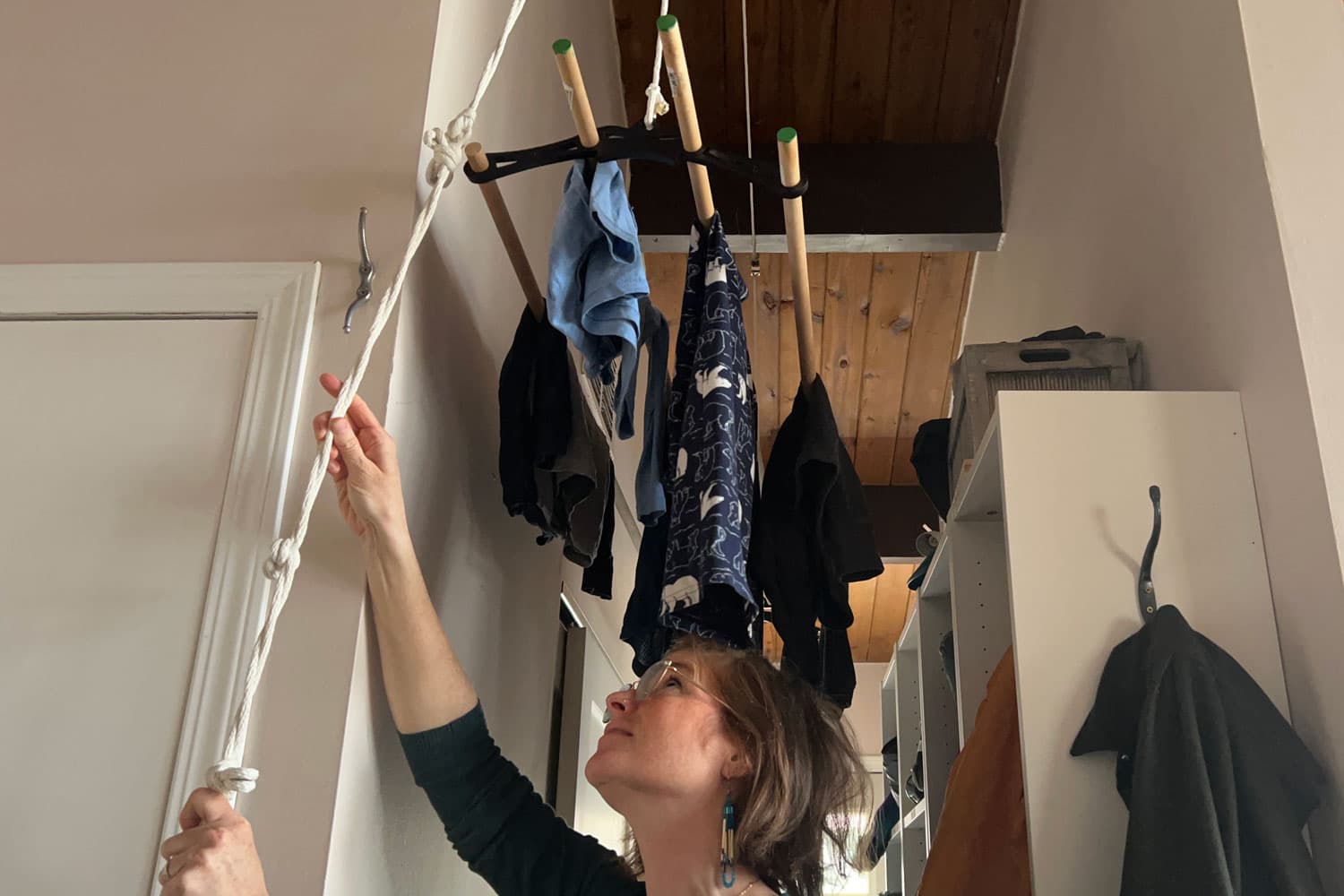 Brianna Sharpe, a fair-skinned woman with brown hair and glasses, pulls on a rope raising a ceiling-mounted drying rack in the hallway of her home.