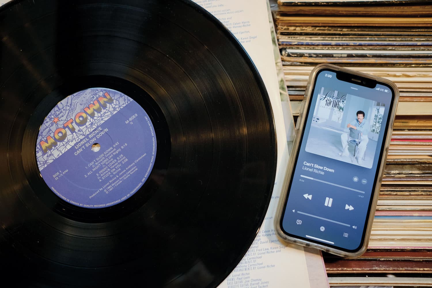 A Lionel Richie vinyl record sitting atop a box filled with record sleeves next to an iPhone streaming the same album.