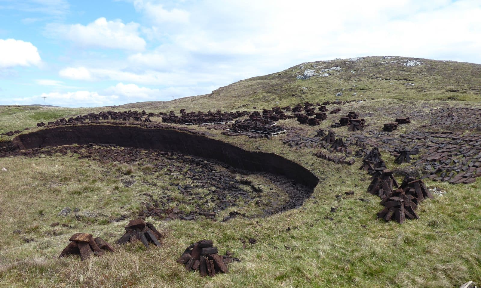Piles of dark peat stacked along the edge of a deep, curved cut in the peat-filled ground. A low hill lies in the background.