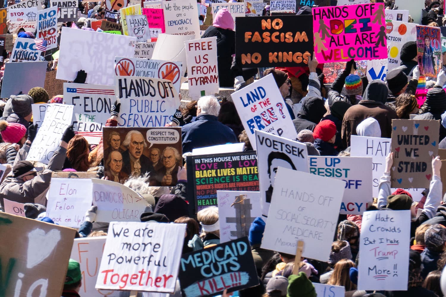 At a rally, protesters hold signs with text like "Hands off DEI," "No fascism no kings," and "Free U.S. from tyranny."