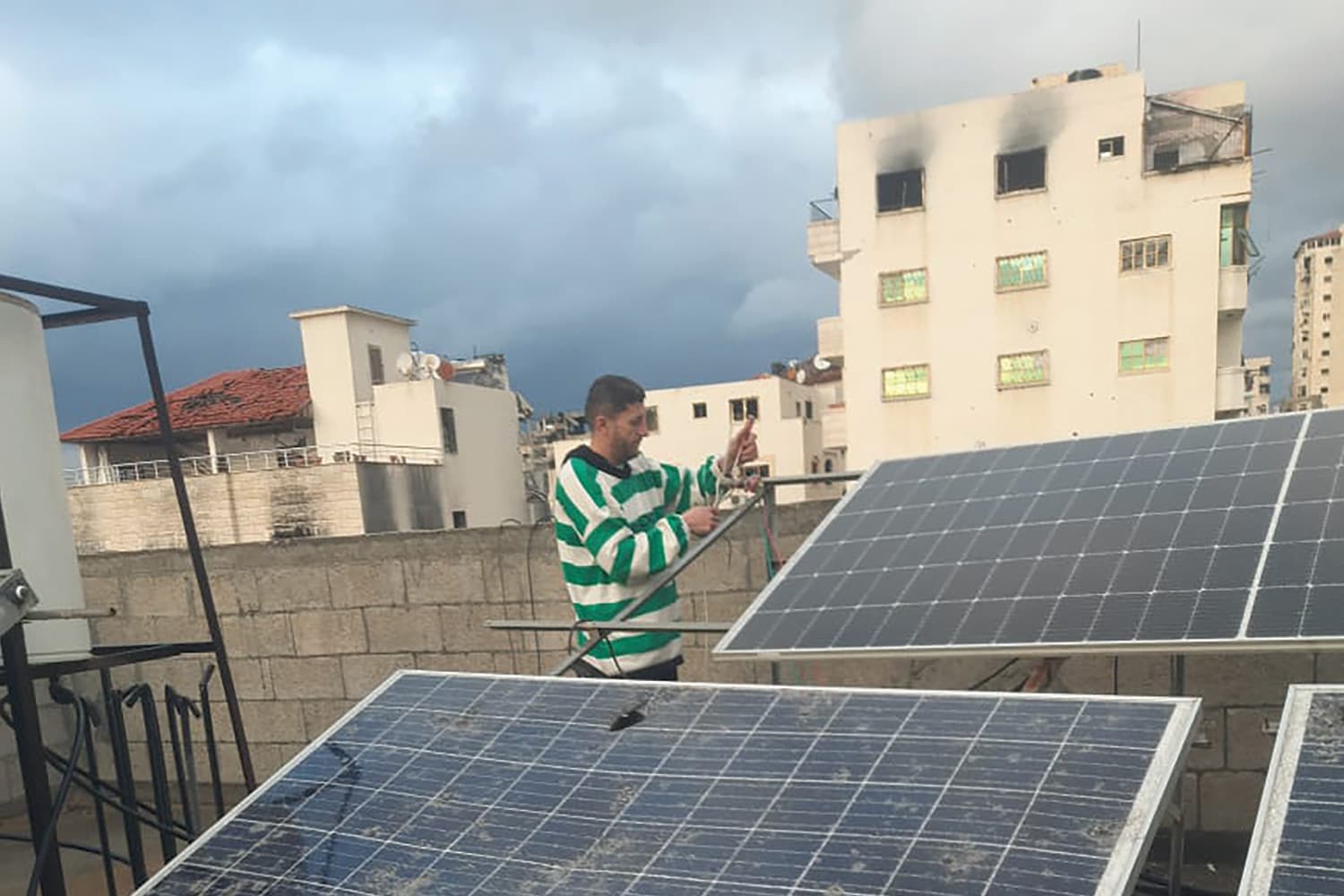 A man wearing a white and green sweater repairs solar panels on a rooftop. Damaged concrete buildings are in the background.